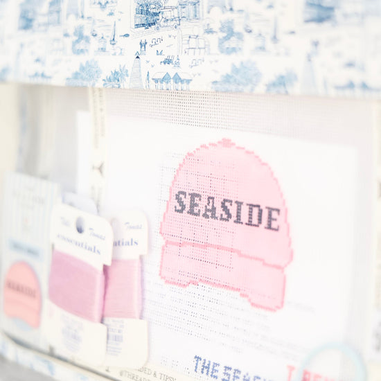 A close-up of a handmade cross-stitch or embroidery sample featuring a pink knit hat with the word 'SEASIDE' stitched across it, surrounded by blue and white patterned fabrics and small fabric swatches.