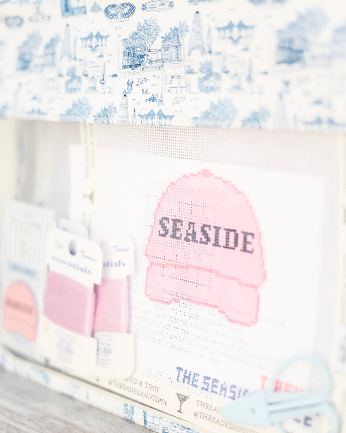A close-up of a handmade cross-stitch or embroidery sample featuring a pink knit hat with the word 'SEASIDE' stitched across it, surrounded by blue and white patterned fabrics and small fabric swatches.