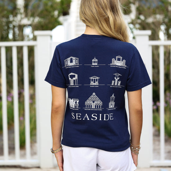 A woman viewed from behind wearing a navy blue t-shirt with a white graphic print of various seaside beach pavilions and the word 'SEASIDE' on the back, paired with white shorts in an outdoor setting.