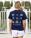 A woman viewed from behind wearing a navy blue t-shirt with a white graphic print of various seaside beach pavilions and the word 'SEASIDE' on the back, paired with white shorts in an outdoor setting.