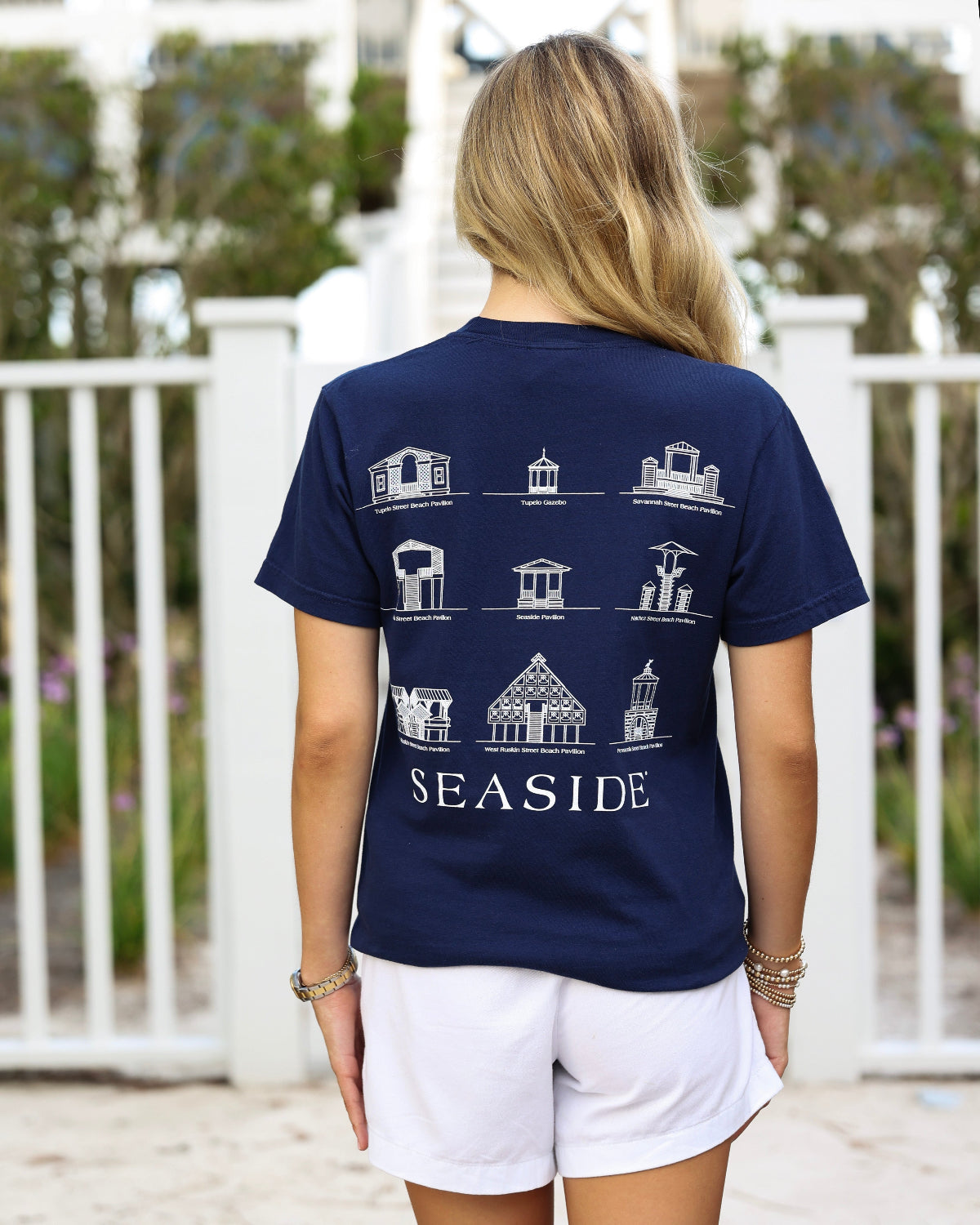 A woman viewed from behind wearing a navy blue t-shirt with a white graphic print of various seaside beach pavilions and the word 'SEASIDE' on the back, paired with white shorts in an outdoor setting.