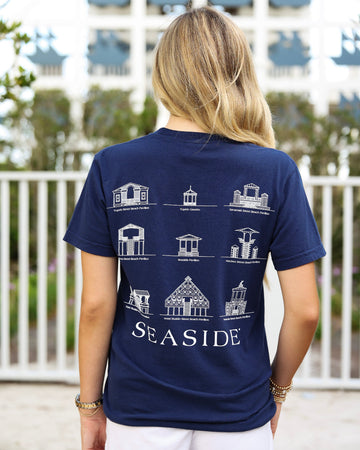 A woman viewed from behind wearing a navy blue t-shirt with white architectural illustrations of various seaside beach pavilions and the word 'SEASIDE' printed on the back, set against an outdoor background with a white fence and a building.