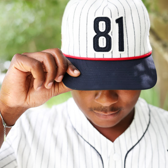 A person wearing a baseball-style cap with a white pinstripe crown, dark navy brim, and the number 81 in black, holding the brim over their eyes. They are also wearing a matching white pinstripe baseball jersey, with a blurred green outdoor background.