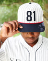 A person wearing a baseball-style cap with a white pinstripe crown, dark navy brim, and the number 81 in black, holding the brim over their eyes. They are also wearing a matching white pinstripe baseball jersey, with a blurred green outdoor background.
