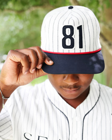 A person wearing a baseball-style cap with a white pinstripe crown, dark navy brim, and the number 81 in black, holding the brim over their eyes. They are also wearing a matching white pinstripe baseball jersey, with a blurred green outdoor background.