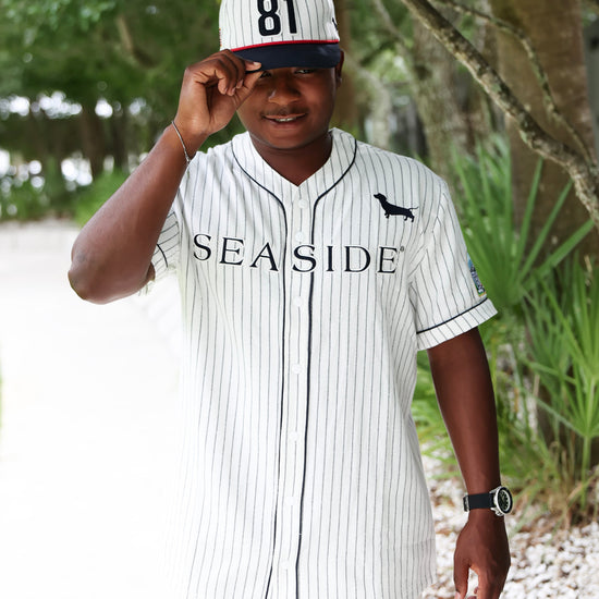 A man wearing a white and navy pinstripe baseball jersey with 'SEASIDE' printed across the chest, a matching cap with '81' on it, and white pants, standing outdoors on a path with trees in the background.