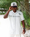 A man wearing a white and navy pinstripe baseball jersey with 'SEASIDE' printed across the chest, a matching cap with '81' on it, and white pants, standing outdoors on a path with trees in the background.