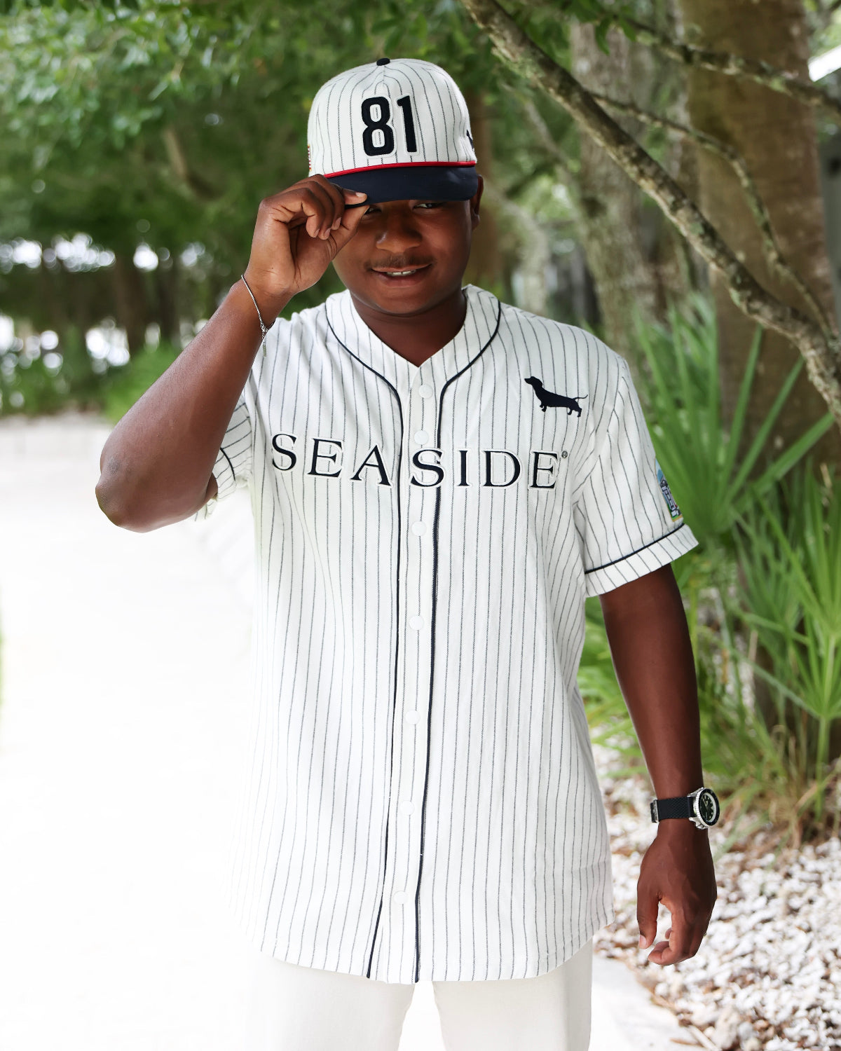 A man wearing a white and navy pinstripe baseball jersey with 'SEASIDE' printed across the chest, a matching cap with '81' on it, and white pants, standing outdoors on a path with trees in the background.