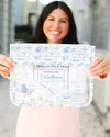 A woman holds up a clear zippered pouch with a blue and white toile-style print border and a central printed insert, shown against a softly blurred outdoor background.