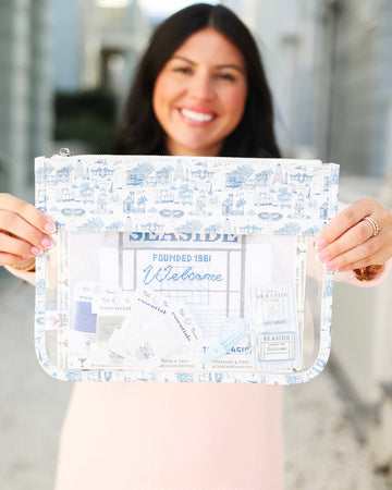 A woman holds up a clear zippered pouch with a blue and white toile-style print border and a central printed insert, shown against a softly blurred outdoor background.