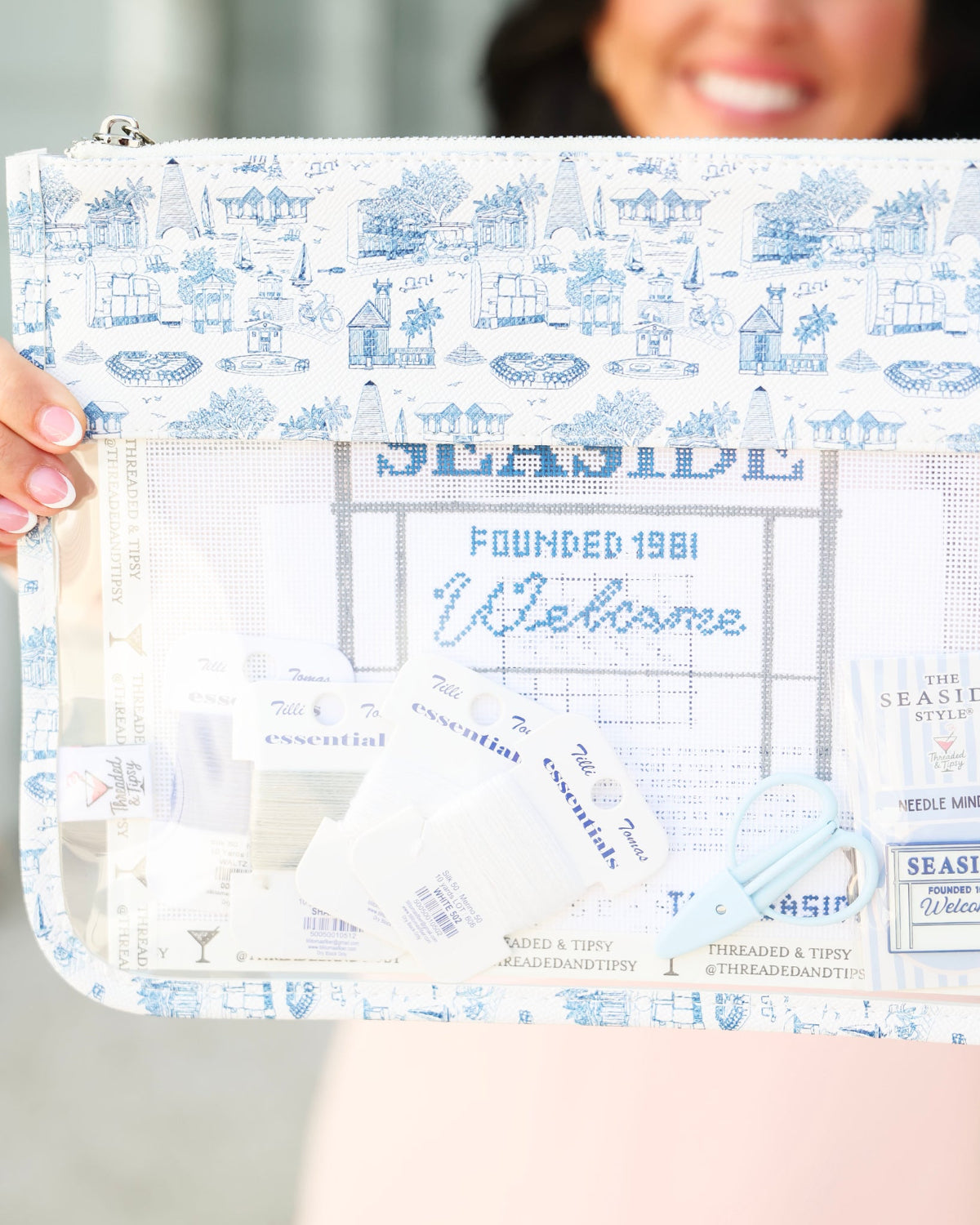 A person holds a clear zip pouch with a blue and white toiletted border. The pouch features a transparent center section displaying various thread and sewing supplies, including spools of thread, a small blue bow, and labels.