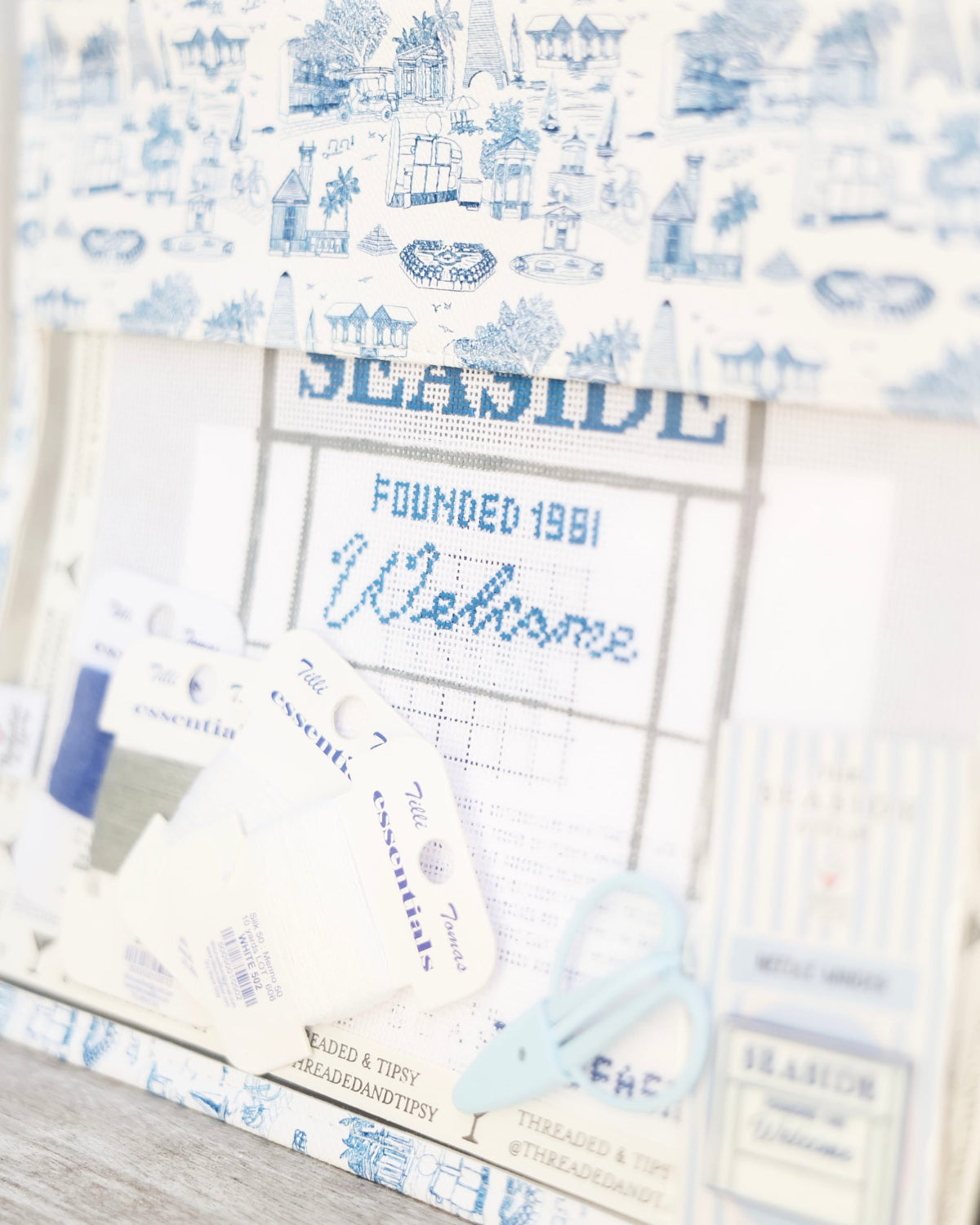 A close-up of a cross-stitch or embroidery fabric design featuring the seaside welcome sign in blue and white. 