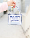 A hand wearing a pink ribbed sleeve holds a small hanging canvas sign that reads 'SEASIDE FOUNDED 1981 Welcome' in blue text on a white background with a navy-and-white rope hanger.