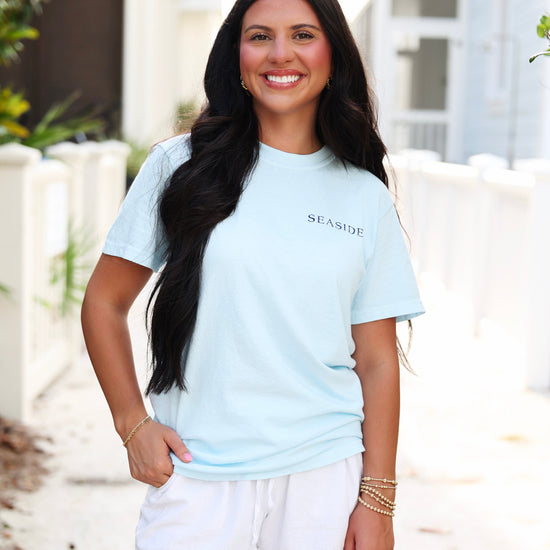 A woman smiling outdoors wearing a light blue short-sleeve t-shirt with 'SEASIDE' printed in black on the chest, paired with white shorts.