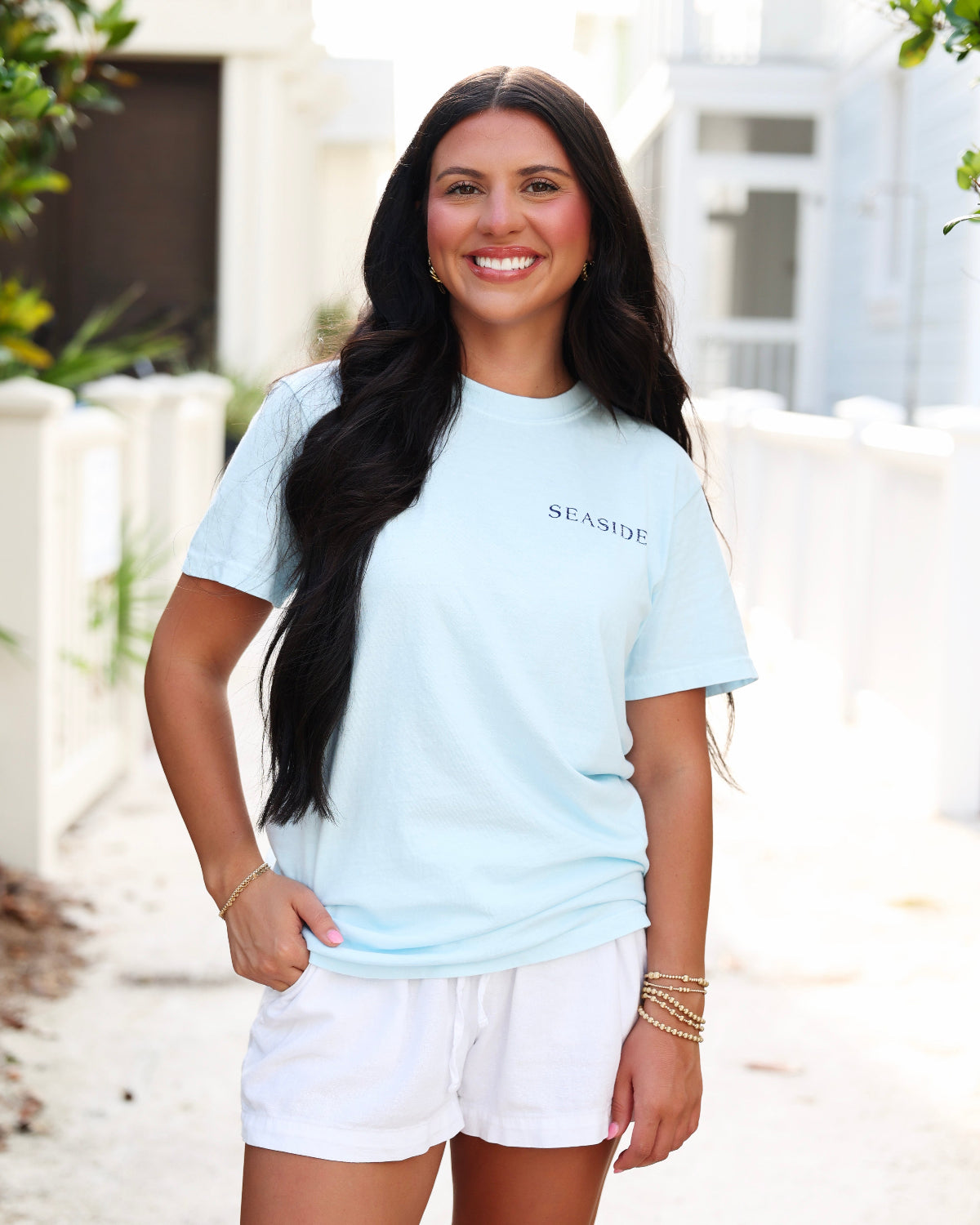 A woman smiling outdoors wearing a light blue short-sleeve t-shirt with 'SEASIDE' printed in black on the chest, paired with white shorts.
