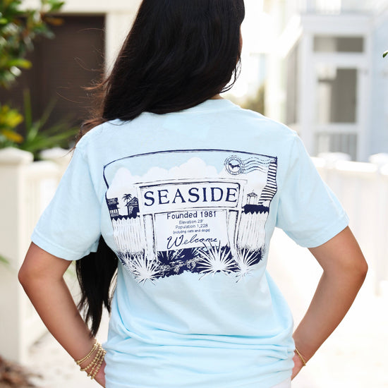 A woman viewed from behind wearing a light blue t-shirt with a large black and white 'SEASIDE' welcome sign graphic on the back, paired with white pants in an outdoor setting.