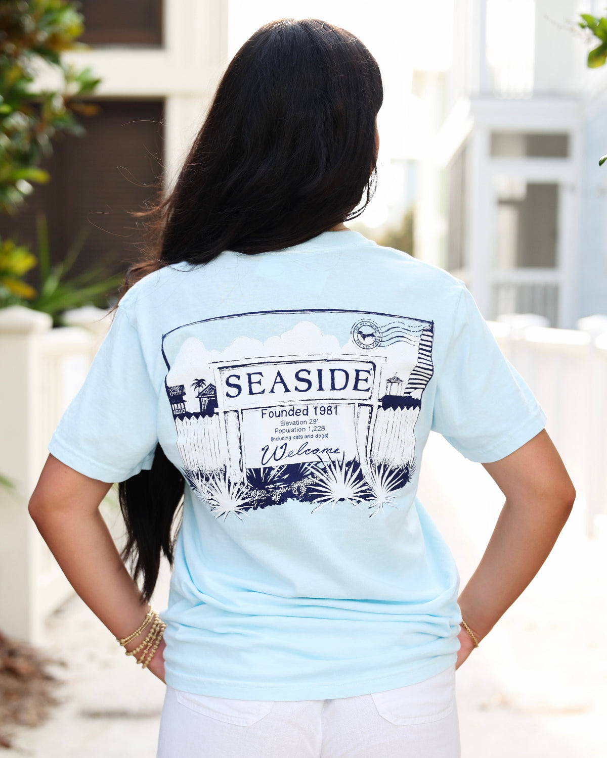 A woman viewed from behind wearing a light blue t-shirt with a large black and white 'SEASIDE' welcome sign graphic on the back, paired with white pants in an outdoor setting.