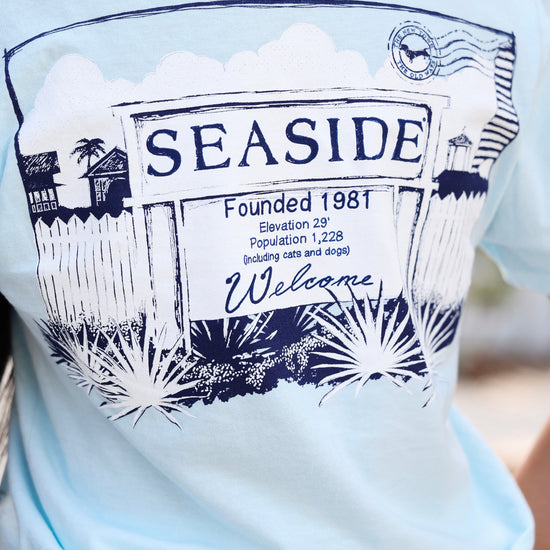 A close-up of a person wearing a light blue t-shirt with a dark navy blue graphic print that reads 'SEASIDE Founded 1981' and 'Welcome' with a postmark illustration.