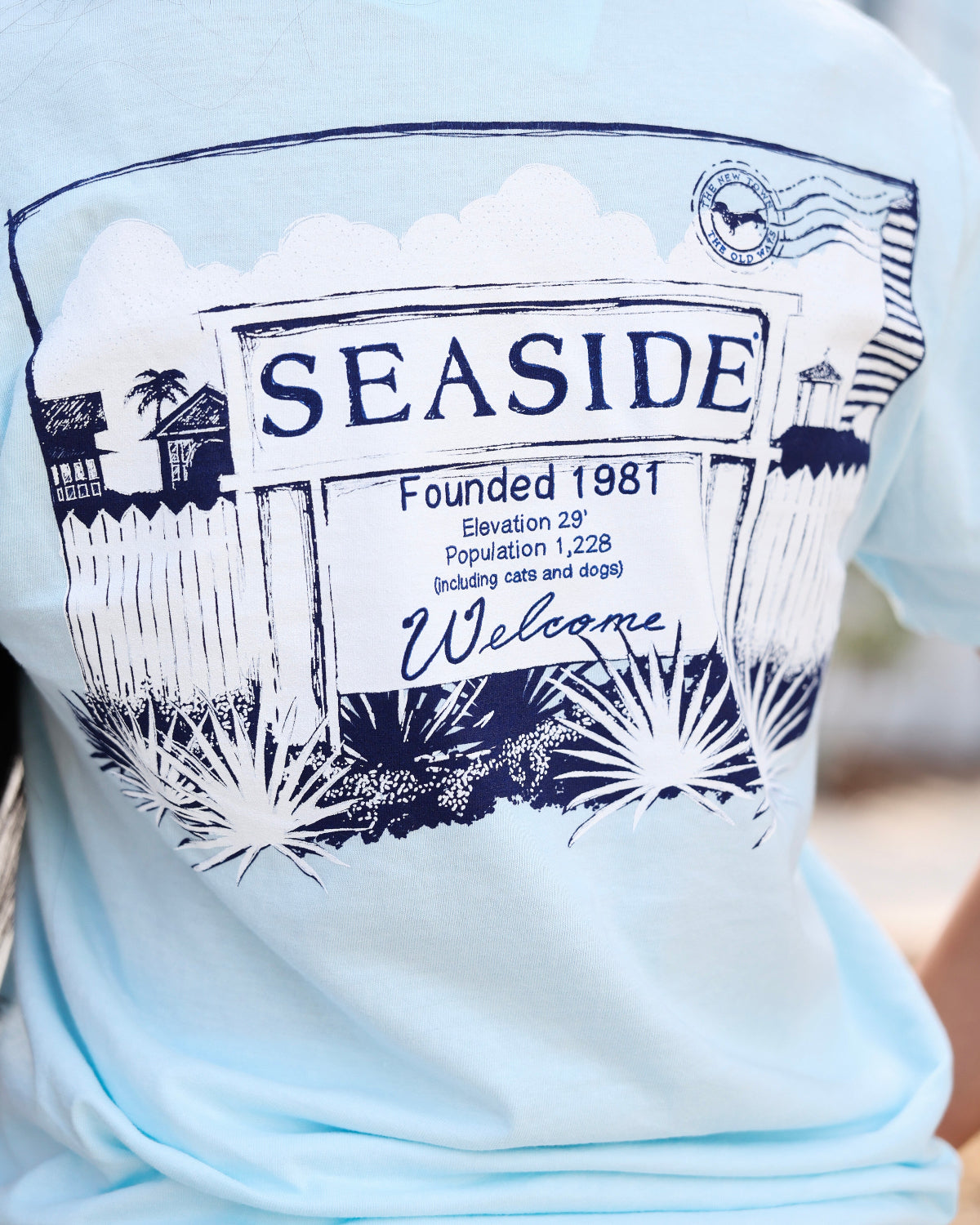 A close-up of a person wearing a light blue t-shirt with a dark navy blue graphic print that reads 'SEASIDE Founded 1981' and 'Welcome' with a postmark illustration.