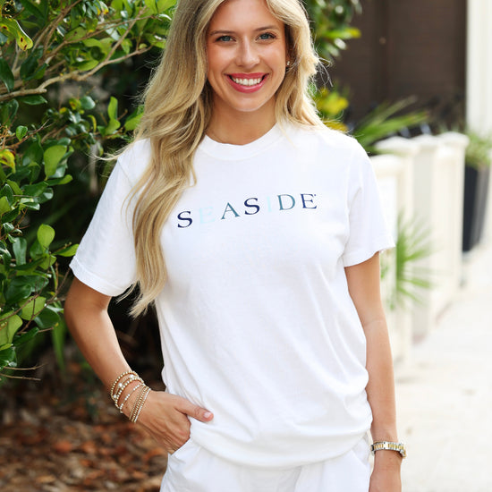 A smiling blonde woman wearing a white short-sleeve t-shirt with 'SEASIDE' printed across the chest in navy and blue lettering, paired with white shorts, standing outdoors in front of green foliage.