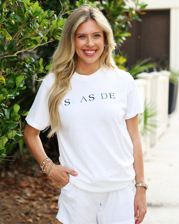 A smiling blonde woman wearing a white short-sleeve t-shirt with 'SEASIDE' printed across the chest in navy and blue lettering, paired with white shorts, standing outdoors in front of green foliage.