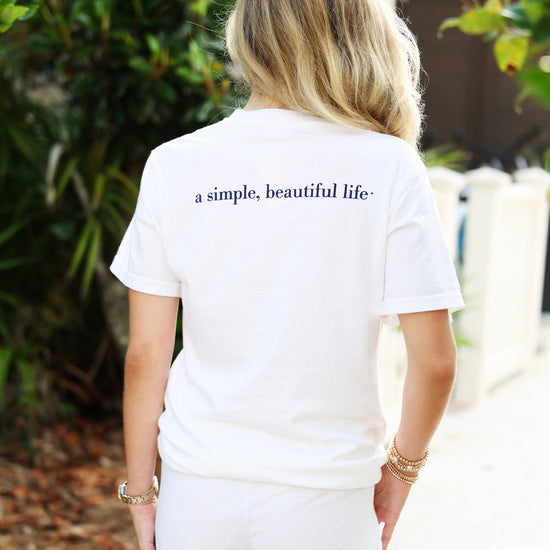 A woman viewed from behind wearing a white short-sleeve T-shirt with the phrase 'a simple, beautiful life.' printed in navy blue across the upper back, paired with white shorts in an outdoor garden setting.