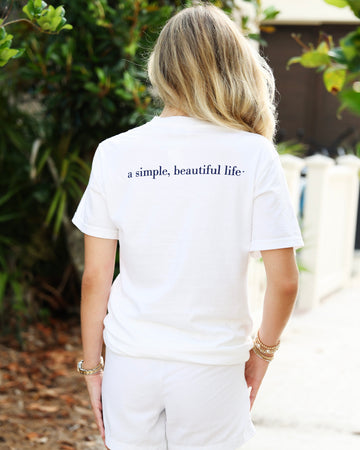 A woman viewed from behind wearing a white short-sleeve T-shirt with the phrase 'a simple, beautiful life.' printed in navy blue across the upper back, paired with white shorts in an outdoor garden setting.