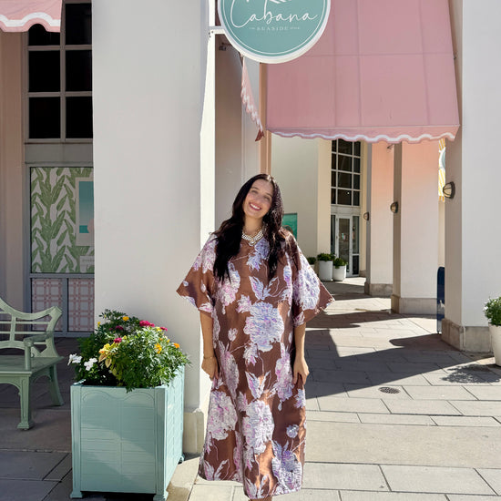 A woman wearing a brown and purple floral midi dress stands on a paved outdoor walkway in front of a building with pink awnings and a round sign that reads 'Cabana'.