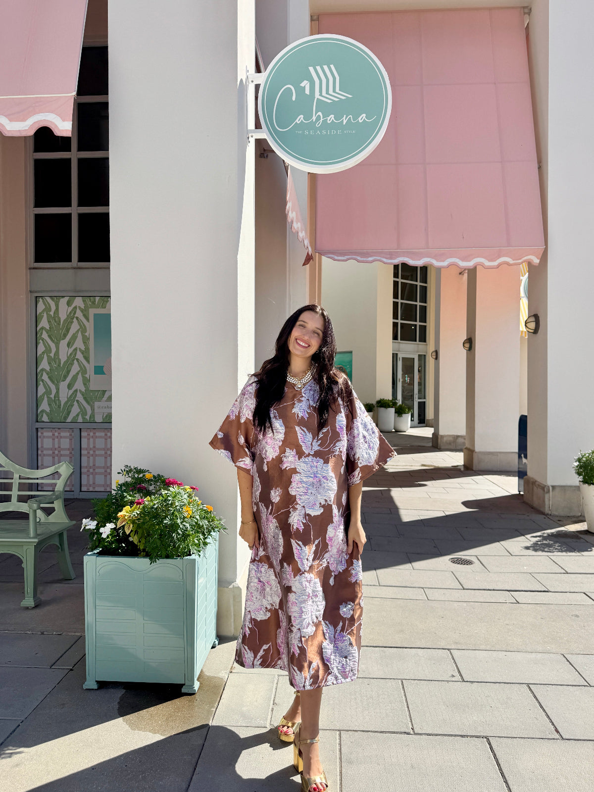 A woman wearing a brown and purple floral midi dress stands on a paved outdoor walkway in front of a building with pink awnings and a round sign that reads 'Cabana'.