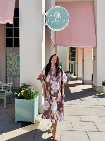 A woman wearing a brown and purple floral midi dress stands on a paved outdoor walkway in front of a building with pink awnings and a round sign that reads 'Cabana'.