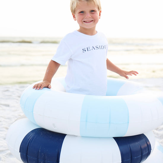 A young blonde boy smiling on a beach while wearing a white t-shirt with 'SEASIDE' printed on the chest, sitting on top of two striped inner tubes (one light blue and white, one navy and white).