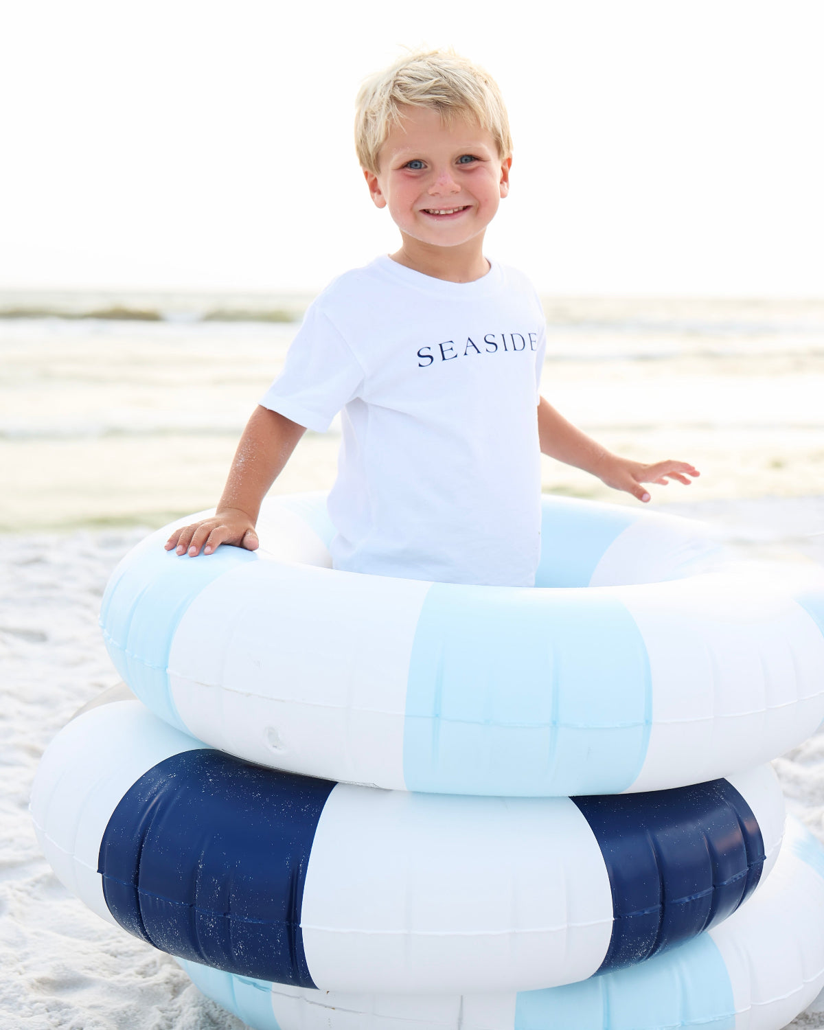 A young blonde boy smiling on a beach while wearing a white t-shirt with 'SEASIDE' printed on the chest, sitting on top of two striped inner tubes (one light blue and white, one navy and white).
