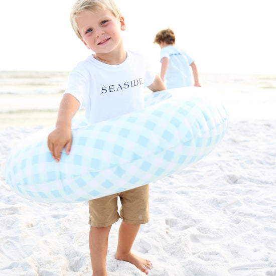 A young boy standing on a sandy beach holds a light blue and white checkered inflatable swim ring. He is wearing a white t-shirt with 'SEASIDE' printed on the front and tan shorts.