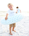 A young boy standing on a sandy beach holds a light blue and white checkered inflatable swim ring. He is wearing a white t-shirt with 'SEASIDE' printed on the front and tan shorts.