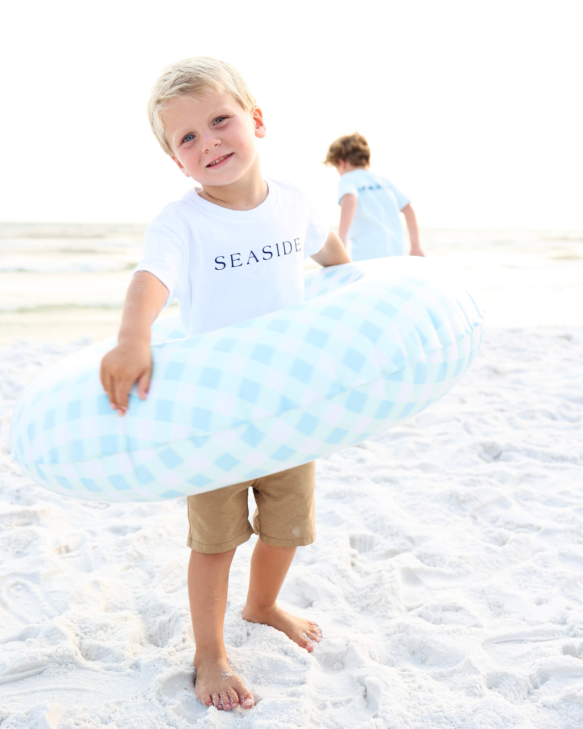 A young boy standing on a sandy beach holds a light blue and white checkered inflatable swim ring. He is wearing a white t-shirt with 'SEASIDE' printed on the front and tan shorts.