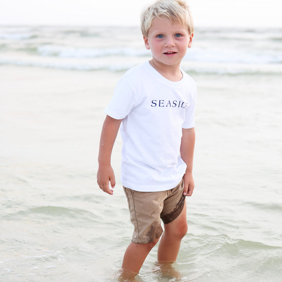A young blond boy standing in shallow ocean water at the beach, wearing a white short-sleeve T-shirt with 'SEASIDE' printed in navy across the chest and tan shorts.