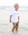 A young blond boy standing in shallow ocean water at the beach, wearing a white short-sleeve T-shirt with 'SEASIDE' printed in navy across the chest and tan shorts.