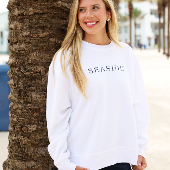 A woman with blonde hair stands outdoors next to a palm tree, wearing a white crewneck sweatshirt with 'SEASIDE' printed in black across the chest and dark shorts.