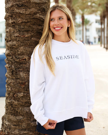 A woman with blonde hair stands outdoors next to a palm tree, wearing a white crewneck sweatshirt with 'SEASIDE' printed in black across the chest and dark shorts.