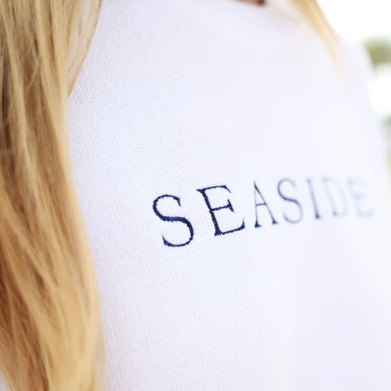A close-up shot of a person wearing a white knit sweater with the word 'SEASIDE' printed in navy blue serif letters across the chest.