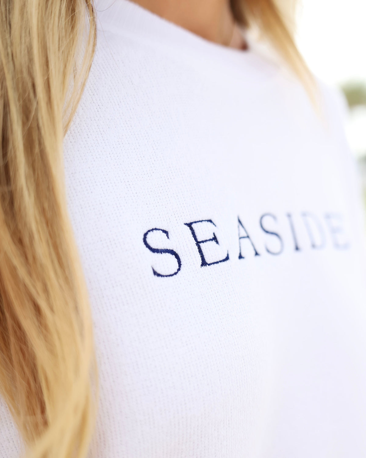 A close-up shot of a person wearing a white knit sweater with the word 'SEASIDE' printed in navy blue serif letters across the chest.