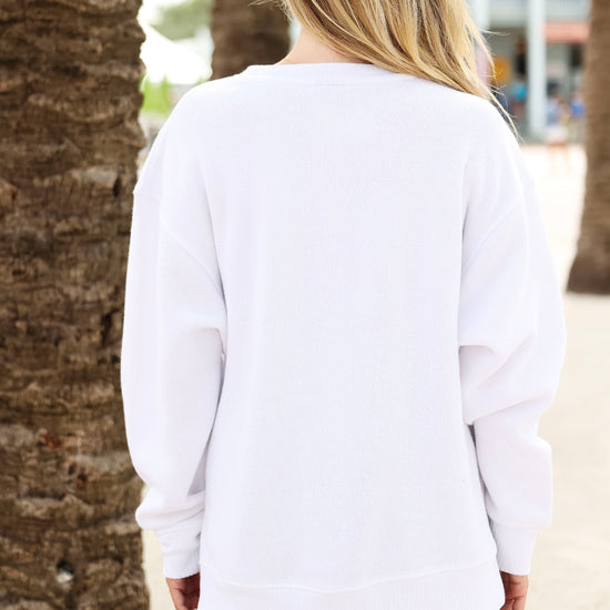 A woman viewed from behind wearing an oversized white long-sleeve sweatshirt and dark shorts, standing outdoors between palm tree trunks.