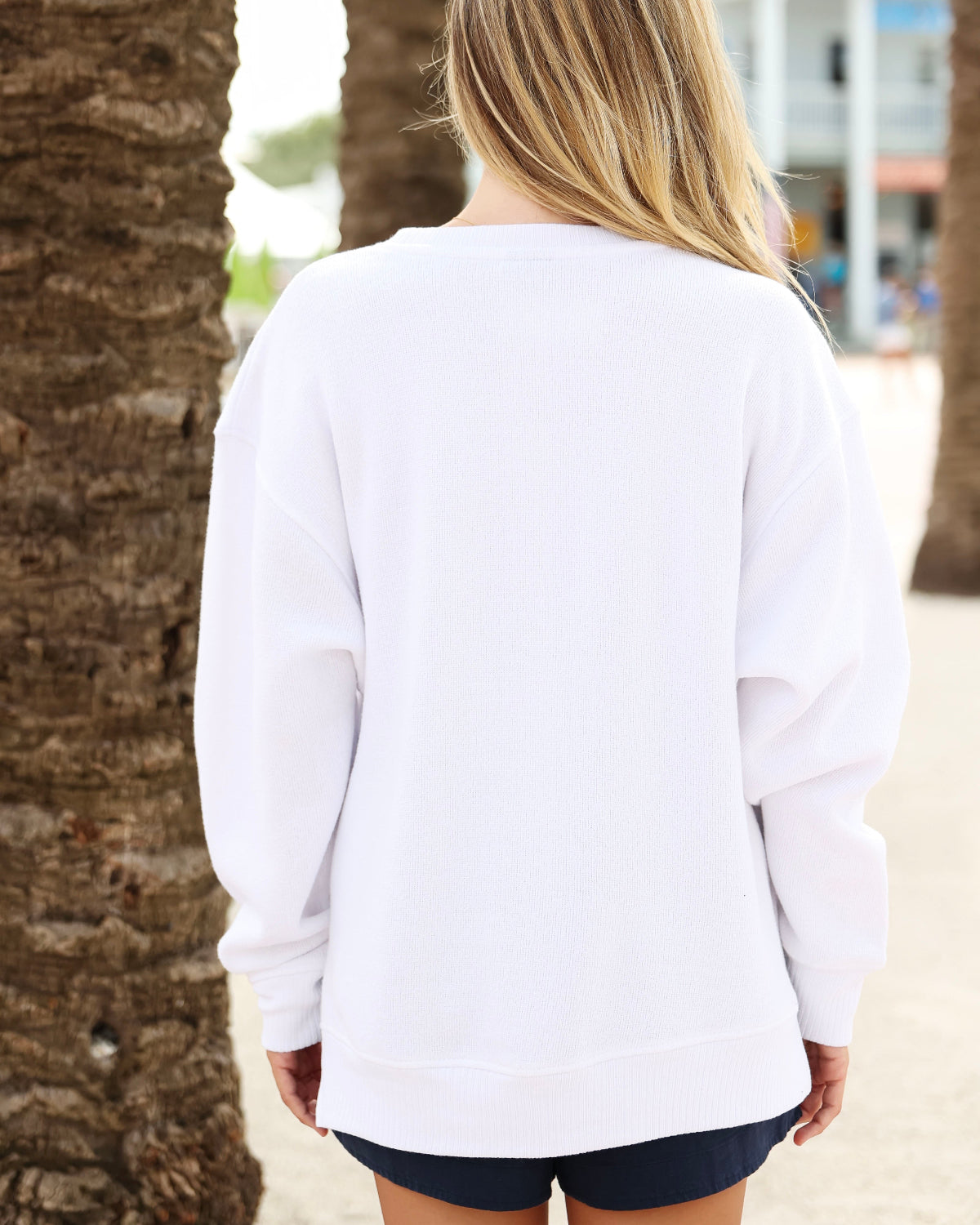 A woman viewed from behind wearing an oversized white long-sleeve sweatshirt and dark shorts, standing outdoors between palm tree trunks.