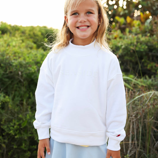 A young girl smiling outdoors wearing a white crewneck sweatshirt and a light blue skirt, with green foliage in the background.