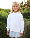 A young girl smiling outdoors wearing a white crewneck sweatshirt and a light blue skirt, with green foliage in the background.