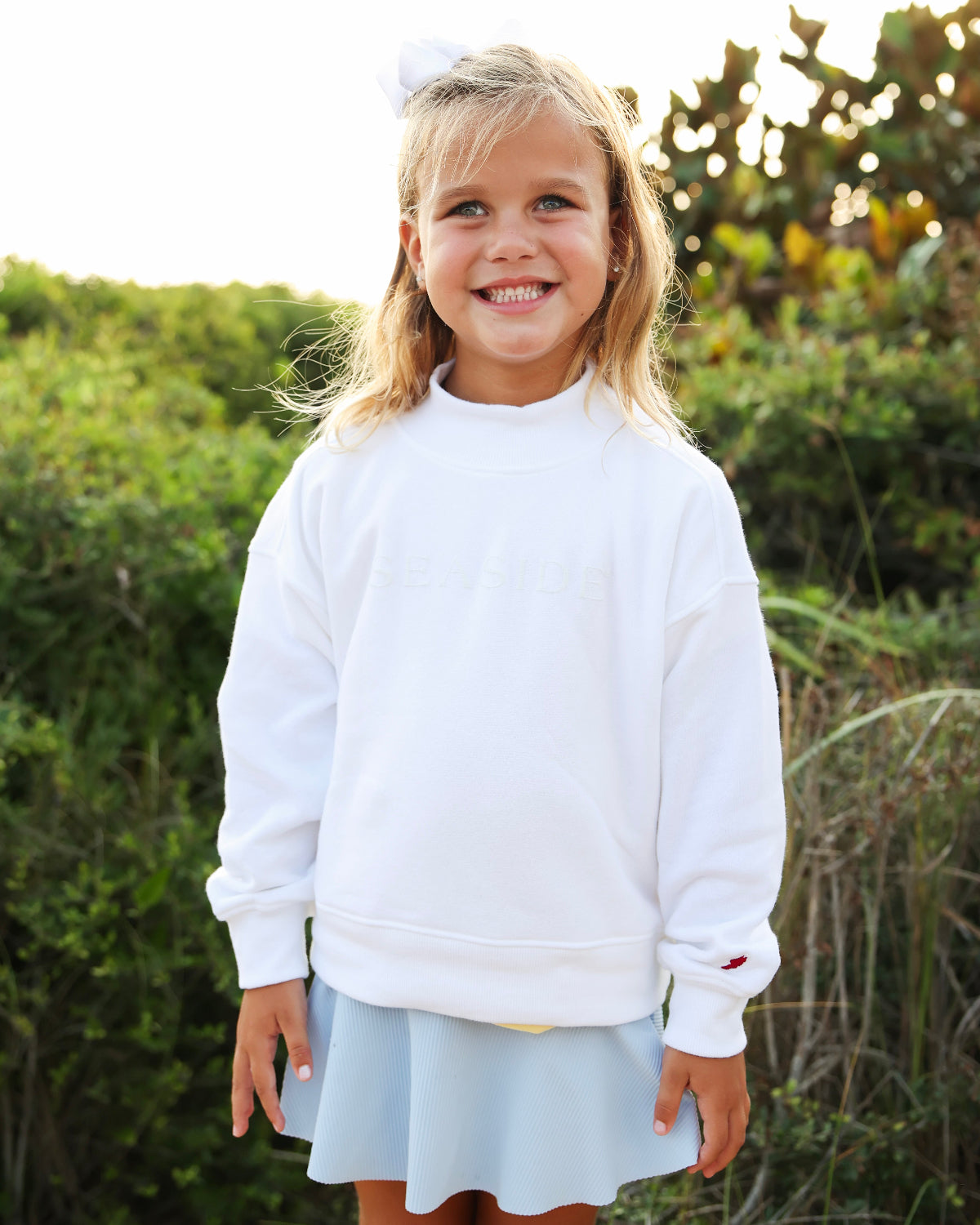 A young girl smiling outdoors wearing a white crewneck sweatshirt and a light blue skirt, with green foliage in the background.