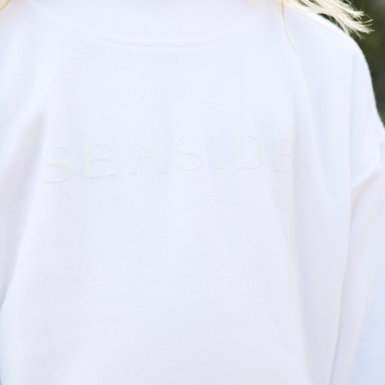 Close-up of a person wearing a white crewneck sweatshirt with the word 'SEASIDE' subtly embroidered across the chest.