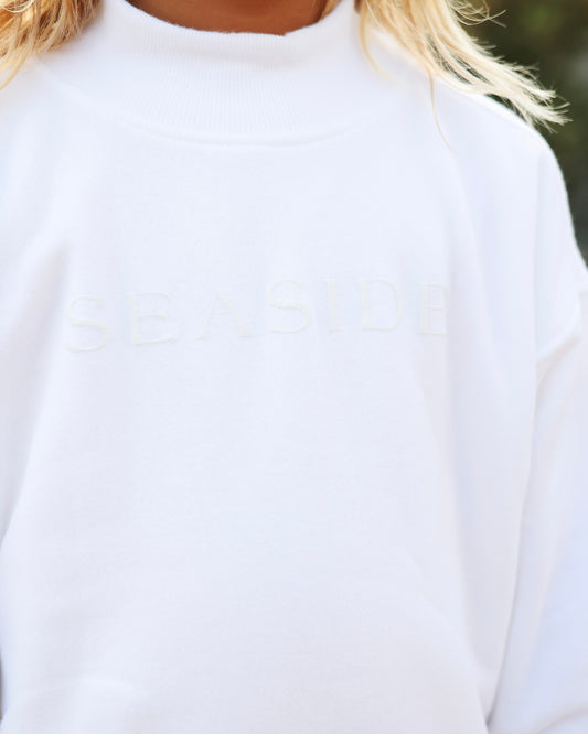 Close-up of a person wearing a white crewneck sweatshirt with the word 'SEASIDE' subtly embroidered across the chest.