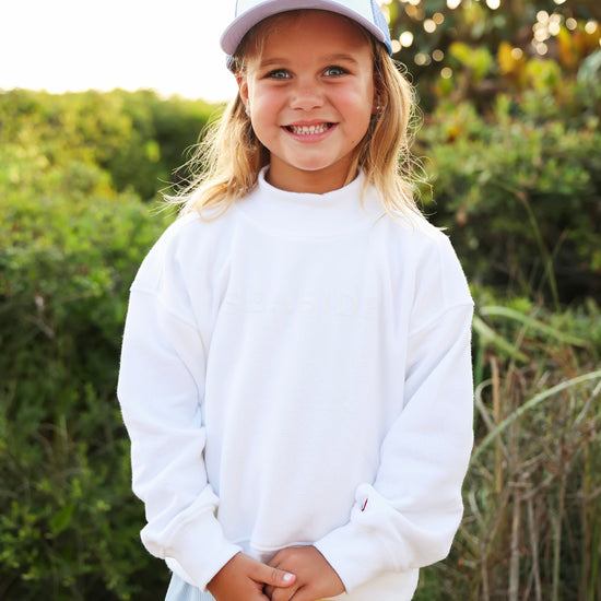A young girl smiling outdoors wearing a white long-sleeve sweatshirt, a light blue skirt, and a white baseball cap with 'SEASIDE' printed on it. The background shows a soft-focus green outdoor setting with warm sunlight.