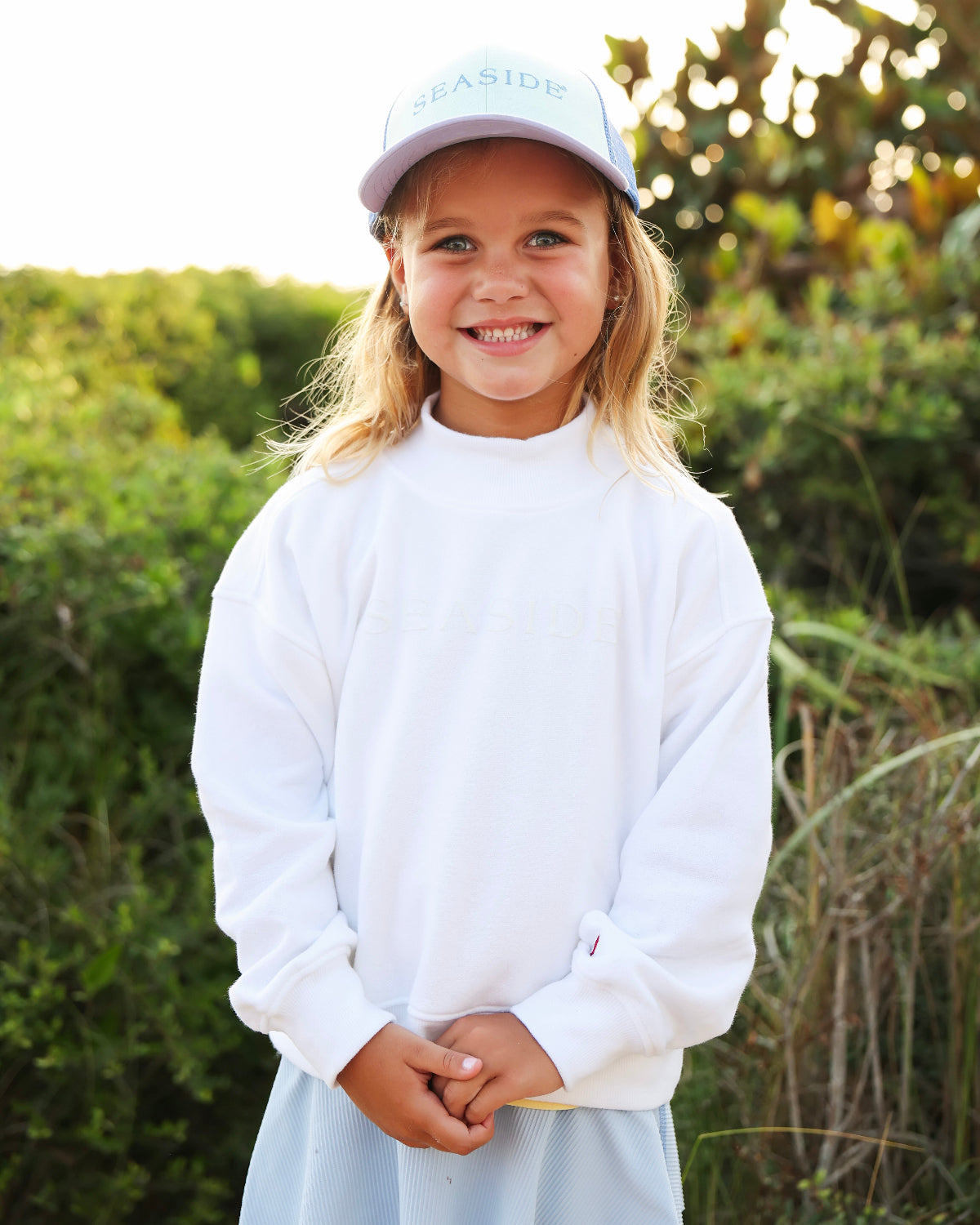 A young girl smiling outdoors wearing a white long-sleeve sweatshirt, a light blue skirt, and a white baseball cap with 'SEASIDE' printed on it. The background shows a soft-focus green outdoor setting with warm sunlight.